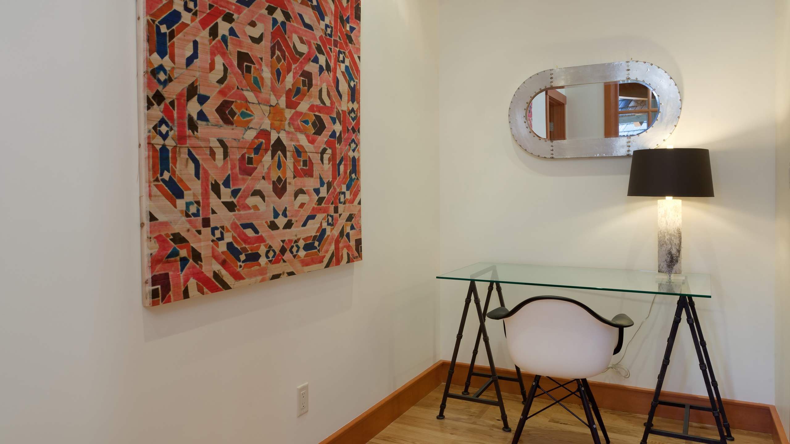 A room with a glass desk, a chair, a lamp, and a large abstract painting on the wall.
