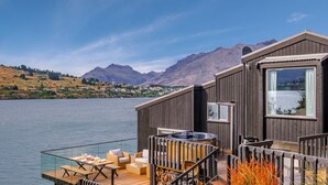 A wooden cabin with a deck overlooking a lake, a hot tub, and a dining area with outdoor furniture. - Queenstown Lakefront Dream | 4 Bed (Queenstown)