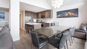 A modern kitchen with a central island, a dining area with a glass table, and a living area with a sofa and a painting of a mountain.
