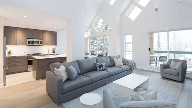 A modern kitchen with dark cabinets and a central island, a grey sofa with cushions, a white coffee table, and a grey armchair.