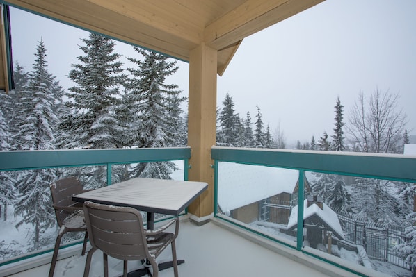 Enjoy a peaceful view of snow-covered trees from this balcony with a table and two chairs.