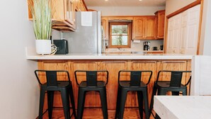 A kitchen with wooden cabinets, a white countertop, a refrigerator, a sink, and three black bar stools. - Lake Cumberland Ohana.  Kid friendly, best views, hot tub. (Burnside)
