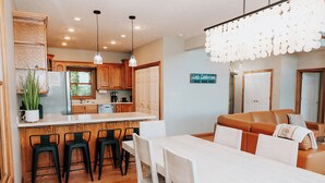 A modern kitchen with wooden cabinets, a white island, and bar stools. There is a dining table with chairs and a sofa in the living area. - Lake Cumberland Ohana.  Kid friendly, best views, hot tub. (Burnside)