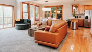 A spacious living room with a leather sofa, a grey armchair, and a coffee table. There is a kitchen area with wooden cabinets and a stainless steel refrigerator. - Lake Cumberland Ohana.  Kid friendly, best views, hot tub. (Burnside)