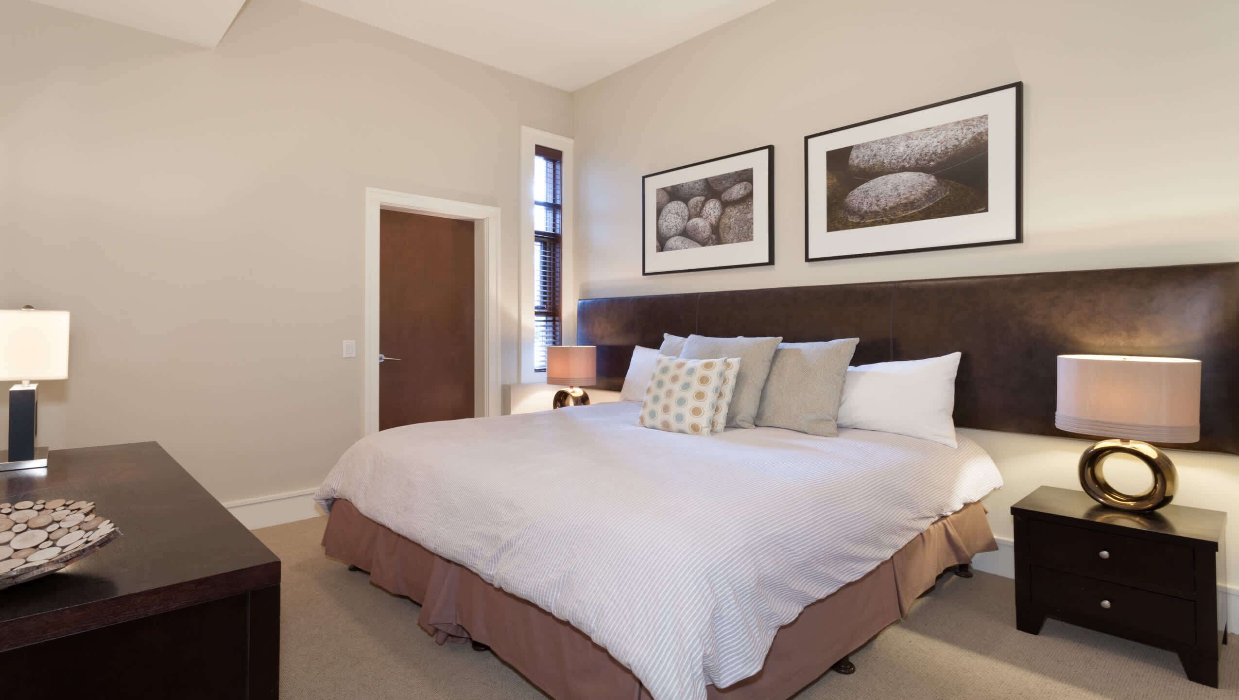 A bedroom with a large bed, two bedside tables, a nightstand, a door, and two framed pictures on the wall.