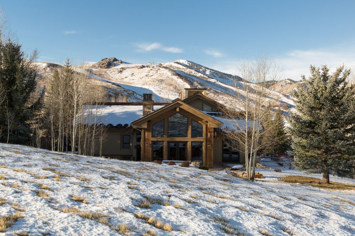Une cabane en bois dotée de grandes fenêtres, entourée de paysages enneigés et d’arbres, avec des montagnes en arrière-plan.
