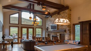 A spacious living room with a pool table, leather chairs, a fireplace, and large windows offering a view of the outdoors. - 2 Miles to Downtown Ketchum | Game Room (Sun Valley)