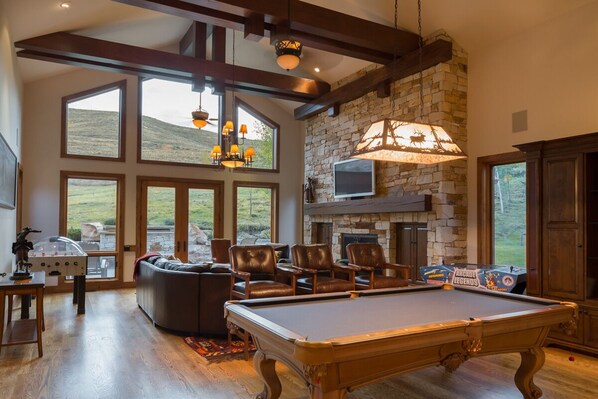 A spacious living room with a pool table, leather chairs, a fireplace, and large windows offering a view of the outdoors. - 2 Miles to Downtown Ketchum | Game Room (Sun Valley)