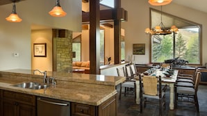 A spacious kitchen with granite countertops, a built-in dishwasher, and a large island. There is a dining area with a table set for six, surrounded by wicker chairs. The room features exposed wooden beams, large windows with a view of trees, and pendant lighting. - 2 Miles to Downtown Ketchum | Game Room (Sun Valley)