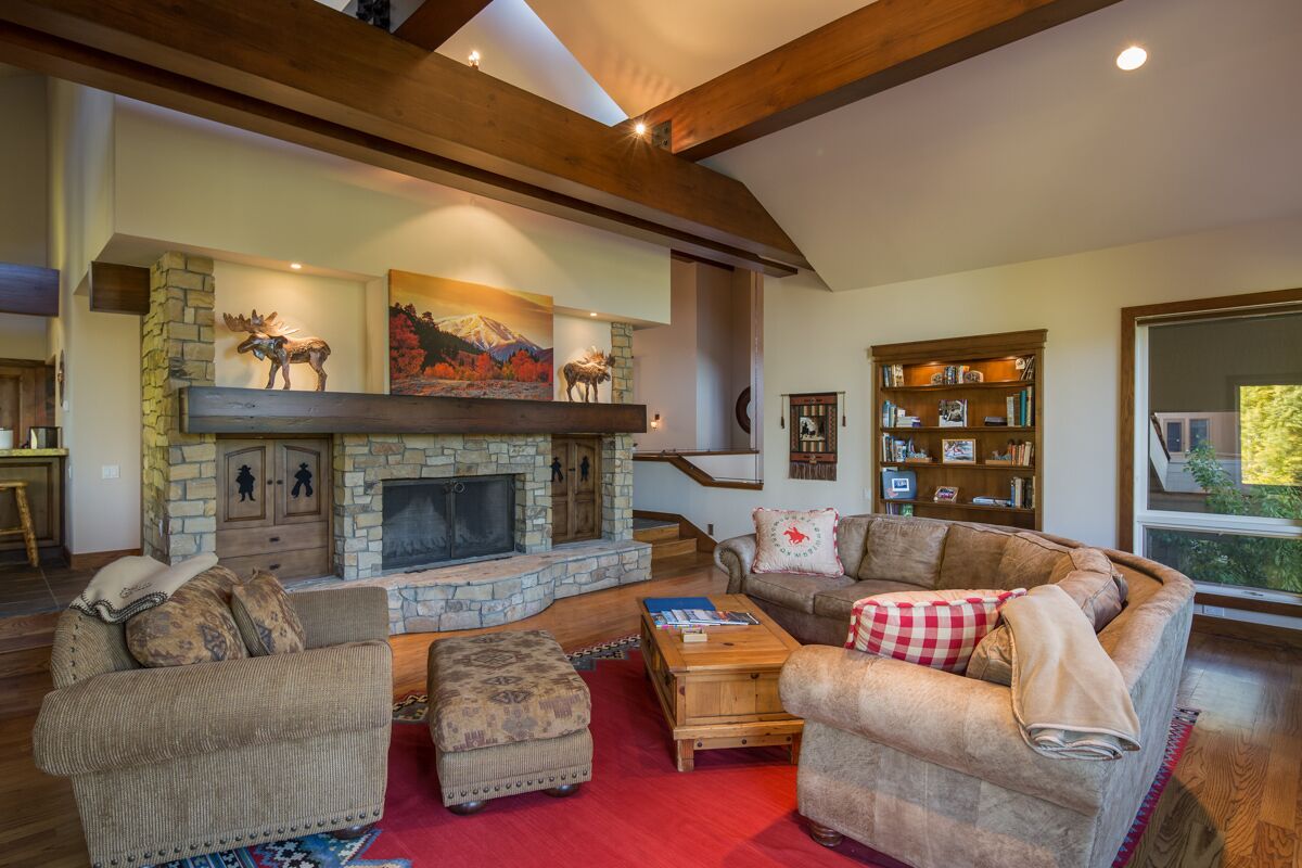 A cozy living room with a stone fireplace, wooden beams, a bookshelf, and comfortable seating.