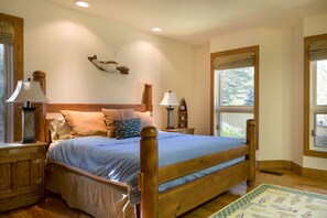 A bedroom with a wooden bed, two bedside tables with lamps, a wall-mounted shelf with decorative items, and a window with a view of greenery. - 2 Miles to Downtown Ketchum | Game Room (Sun Valley)