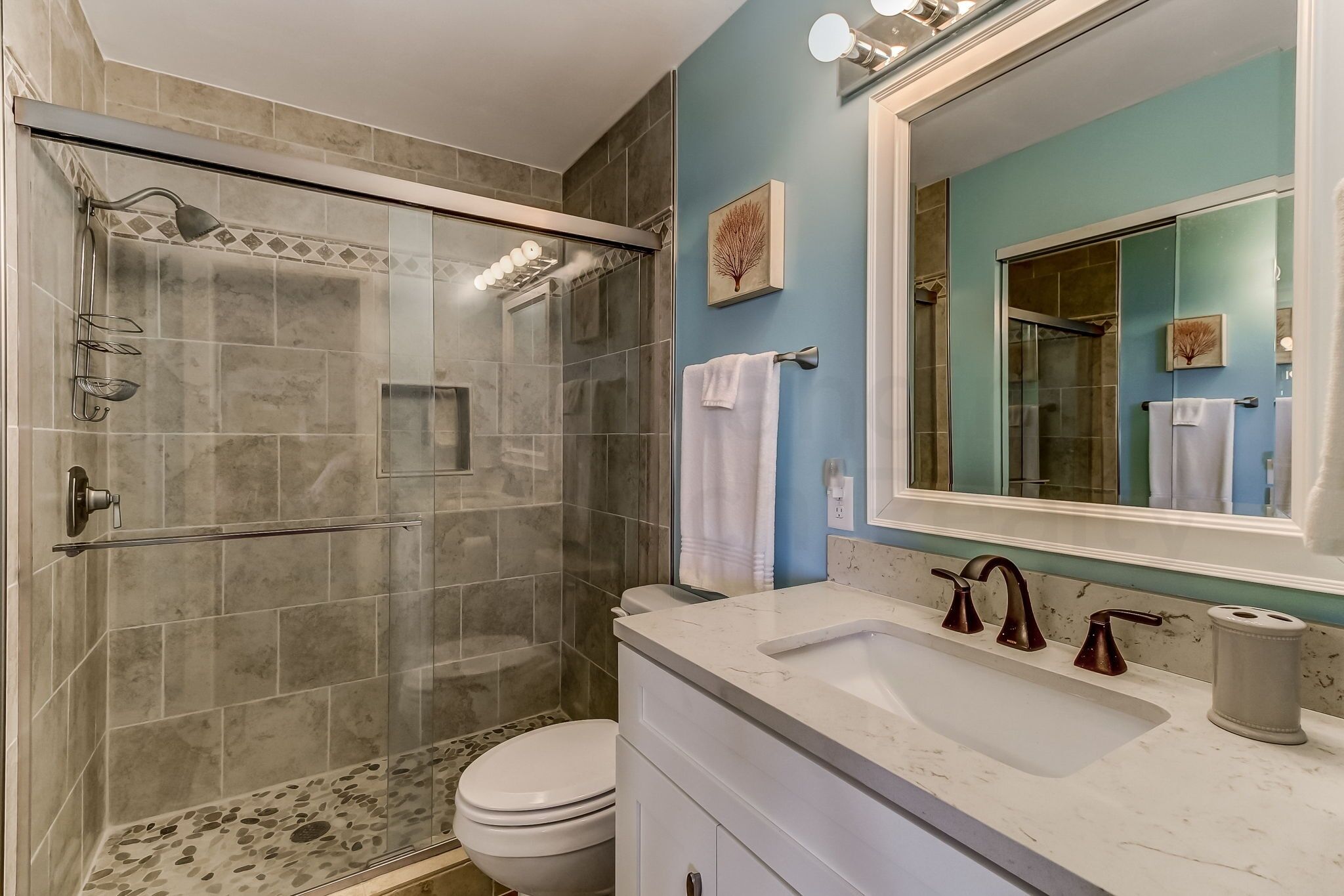Ein Badezimmer mit einer Duschkabine, einer weißen Toilette, einem Waschbecken mit Marmorplatte und einem großen Spiegel mit zwei gerahmten Bildern an der Wand.