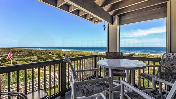 Ein überdachter Außenbereich mit Tisch und Stühlen, mit Blick auf einen Strand und das Meer.