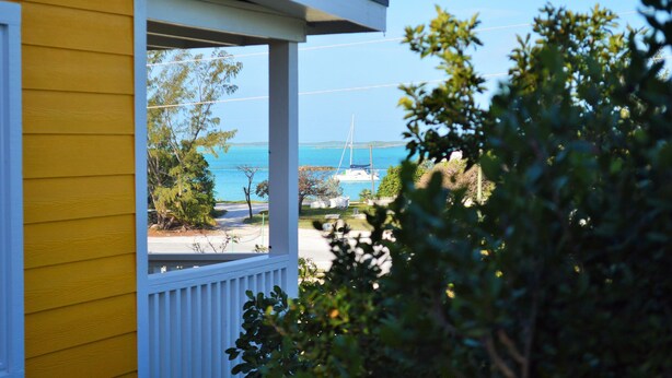 A yellow building with a white porch overlooking a beach with a sailboat. - Cozy Cottage Perfect for 2 (Exuma)