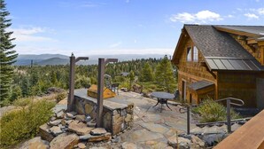 A wooden cabin with a stone patio, outdoor seating, and a view of mountains and forest. - Glacier Luxury Lodge 360 Views (Truckee)
