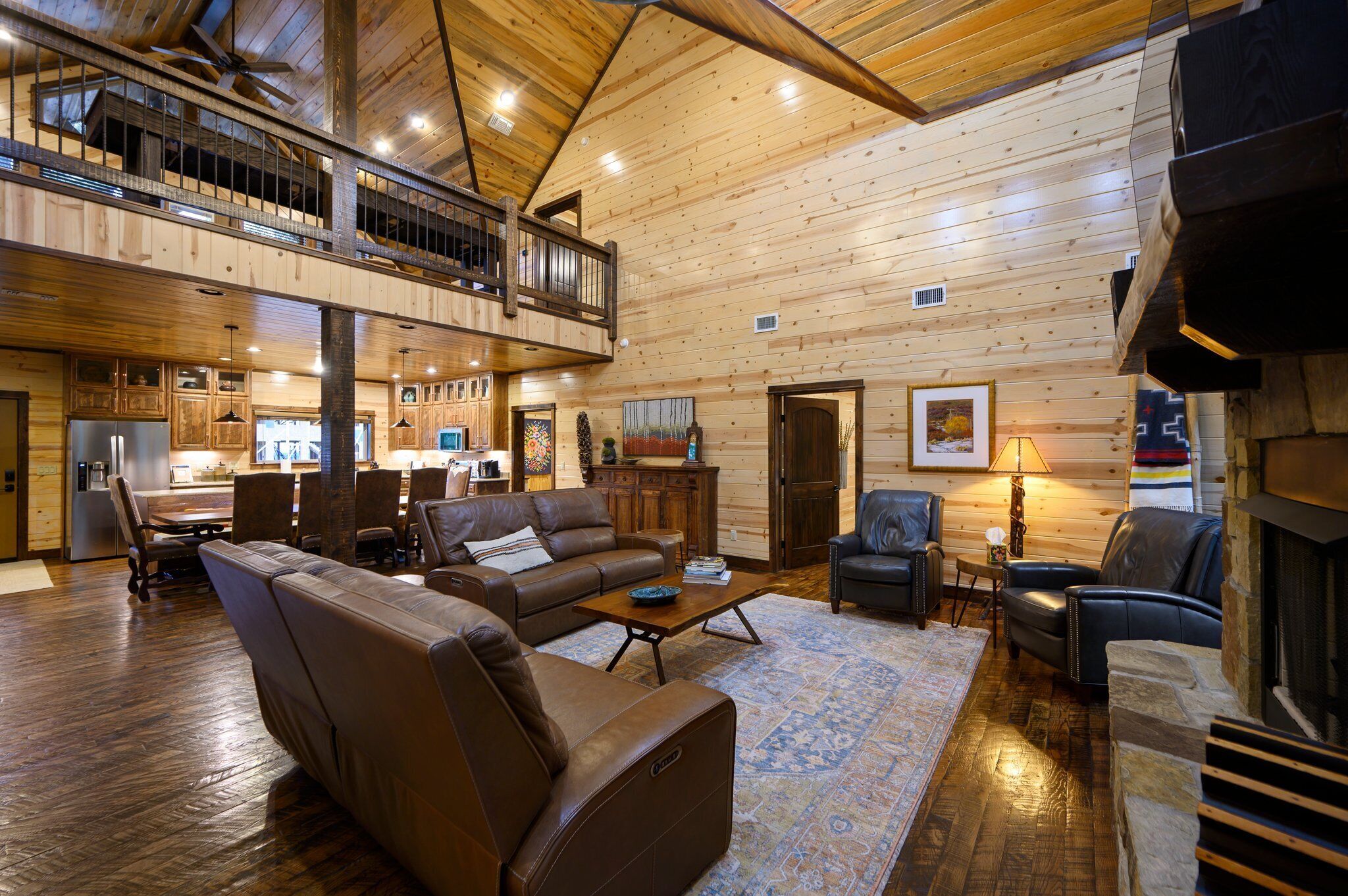 A spacious log cabin interior with a dining area, a living room with leather sofas, a fireplace, and a kitchen visible on the upper level.