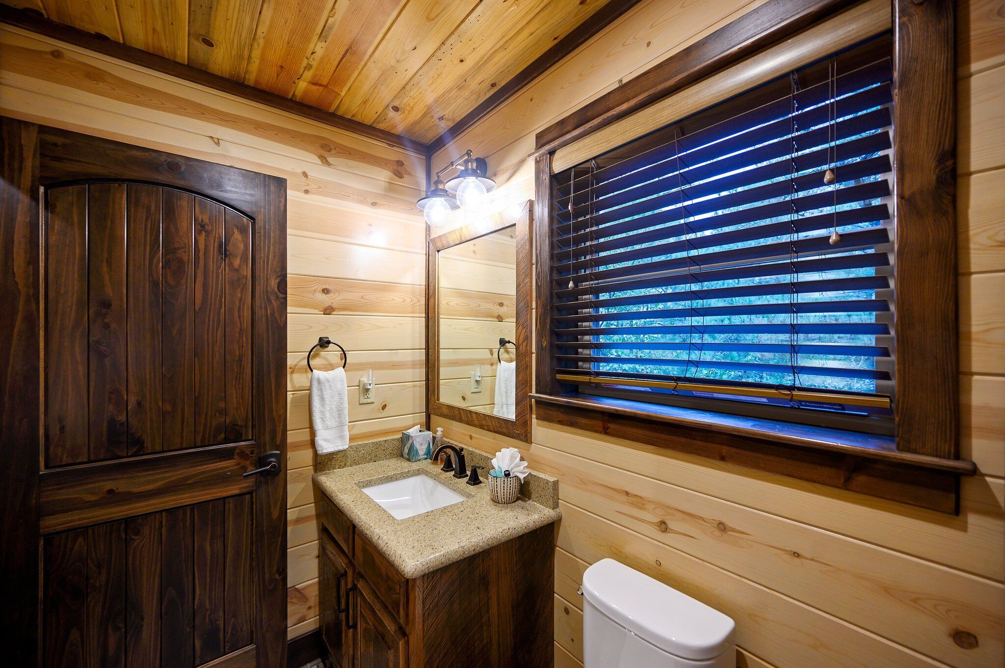 Une salle de bain rustique, aménagée dans une cabane en bois, avec un lavabo, un miroir et une fenêtre équipée de stores.