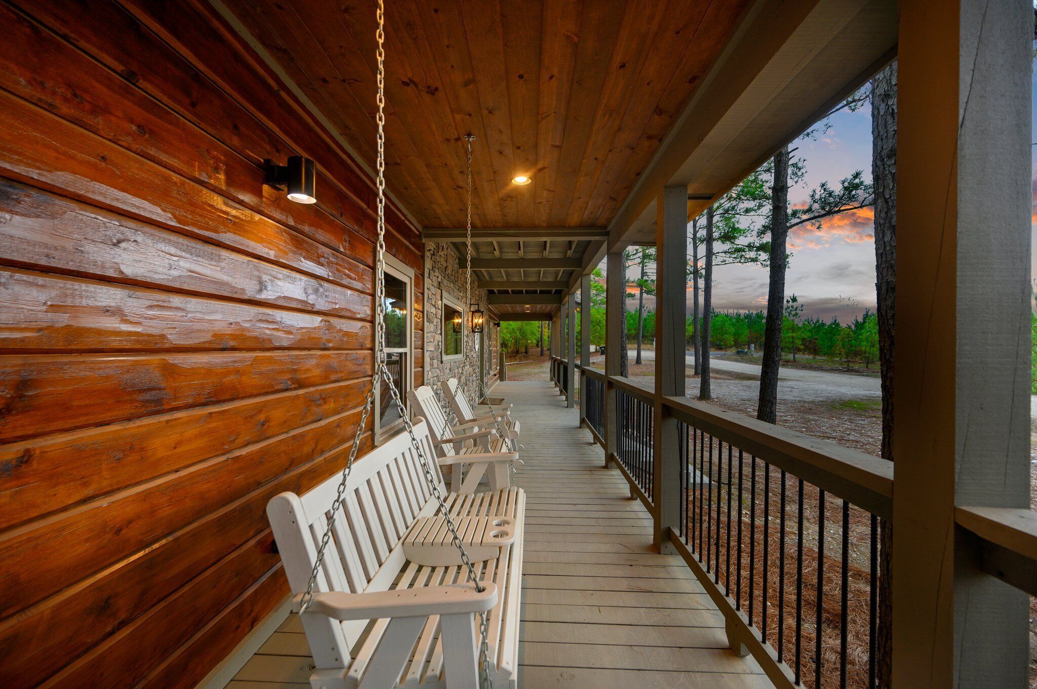 A wooden porch with benches and a railing, overlooking a road and trees.