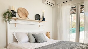 Un dormitorio ordenado con cabecera blanca, cama con ropa de cama blanca y gris, mesita de noche y ventana con vistas al exterior.