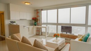 Un salon moderne avec un canapé, une table basse et un téléviseur. Il y a également un coin cuisine avec des placards et une fenêtre agrémentée de rideaux.