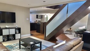 Una sala de estar moderna con un sofá de cuero, una mesa de centro y un televisor sobre un soporte negro. Una escalera conduce a una cocina con microondas.