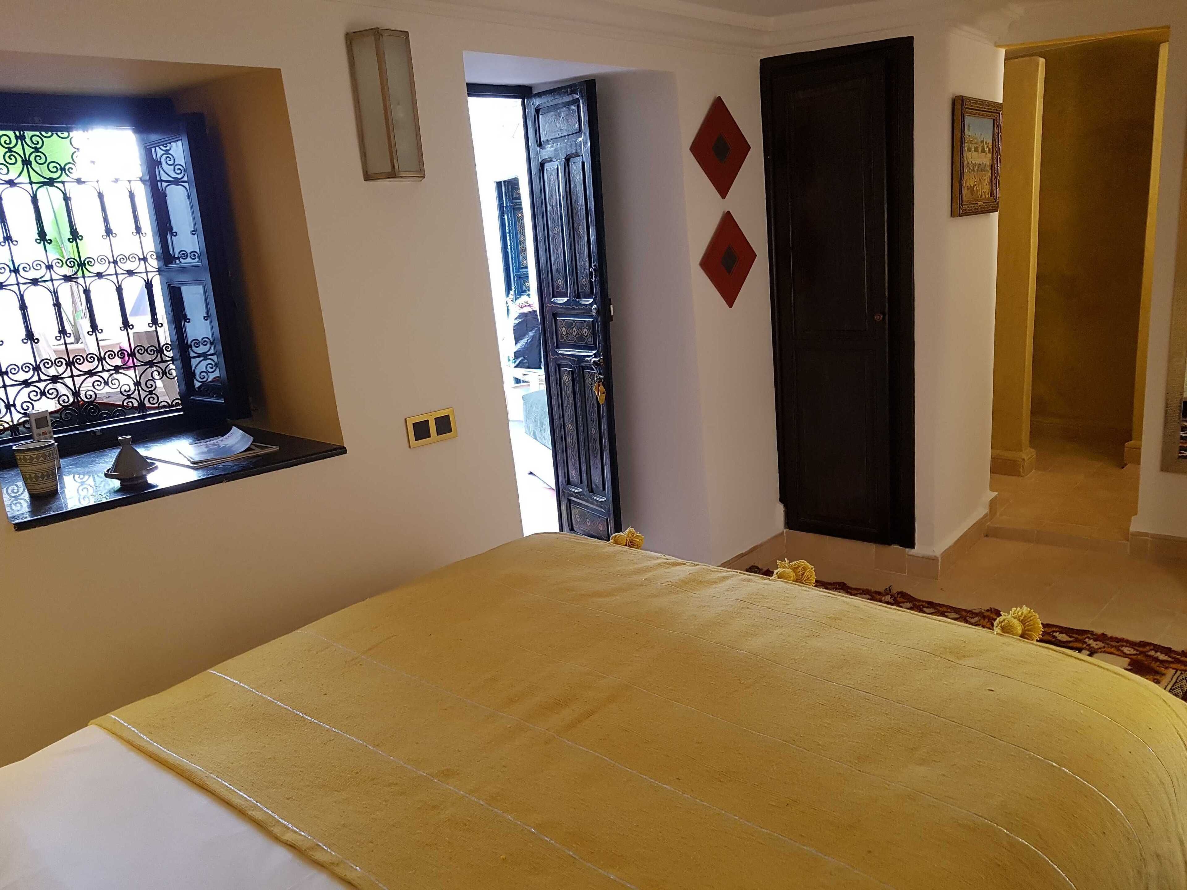 Un dormitori amb llit, finestra amb baranes de ferro decoratives, porta i taula petita amb una tassa i objecte decoratiu.