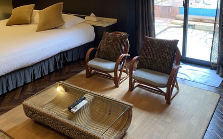 A hotel room with a bed, two wicker chairs, a small table with a remote control, and a view of an outdoor area through a sliding glass door.