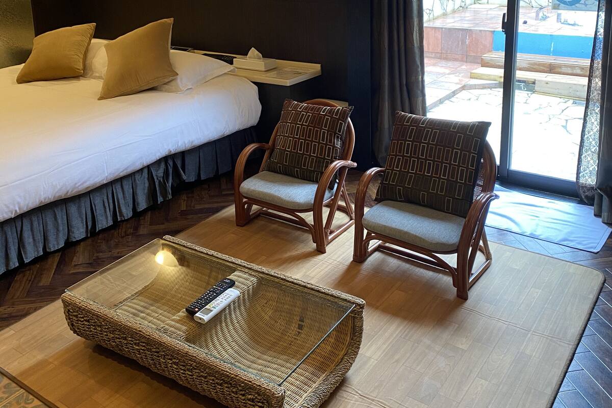 Hotellihuoneessa on sänky, kaksi rottinkituolia, pieni pöytä, jossa on kaukosäädin, ja liukuovesta avautuu näkymä ulkoalueelle.