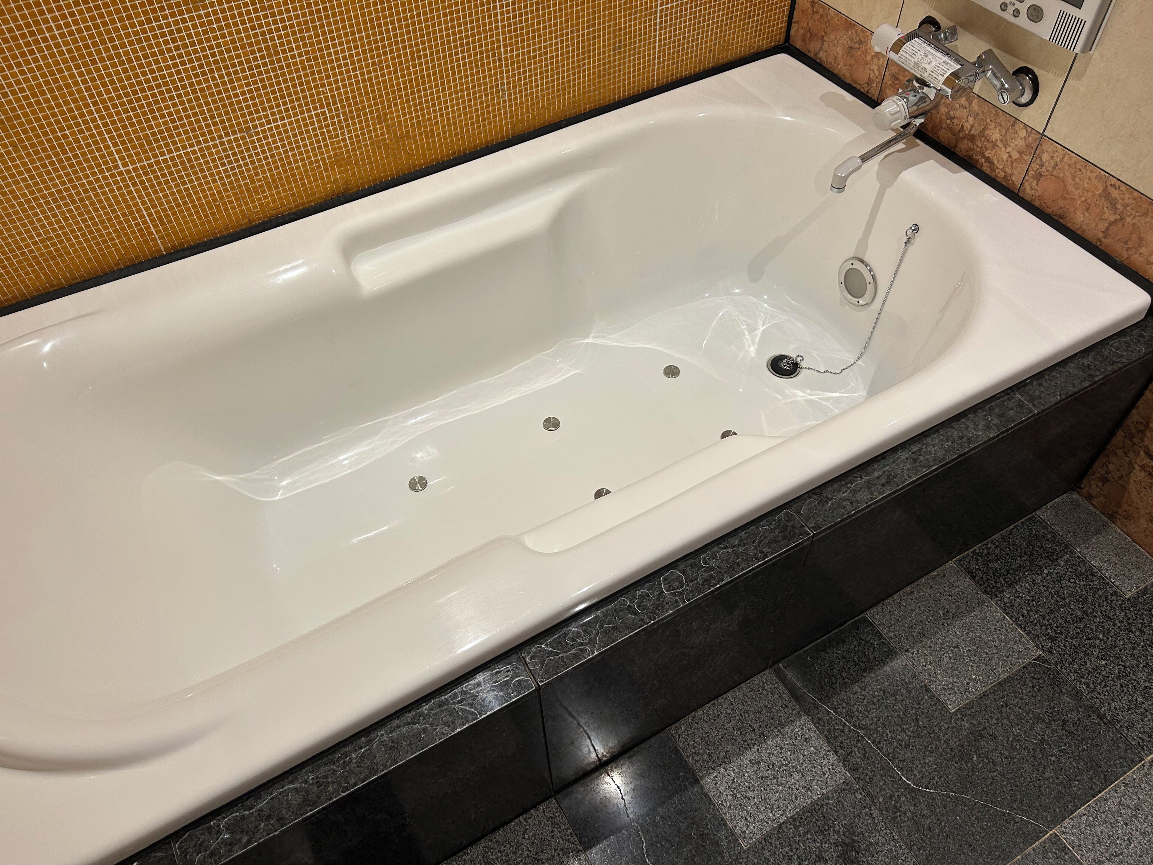 A white bathtub with a single faucet and a drain, set in a bathroom with dark marble tiles and a yellow patterned wall.