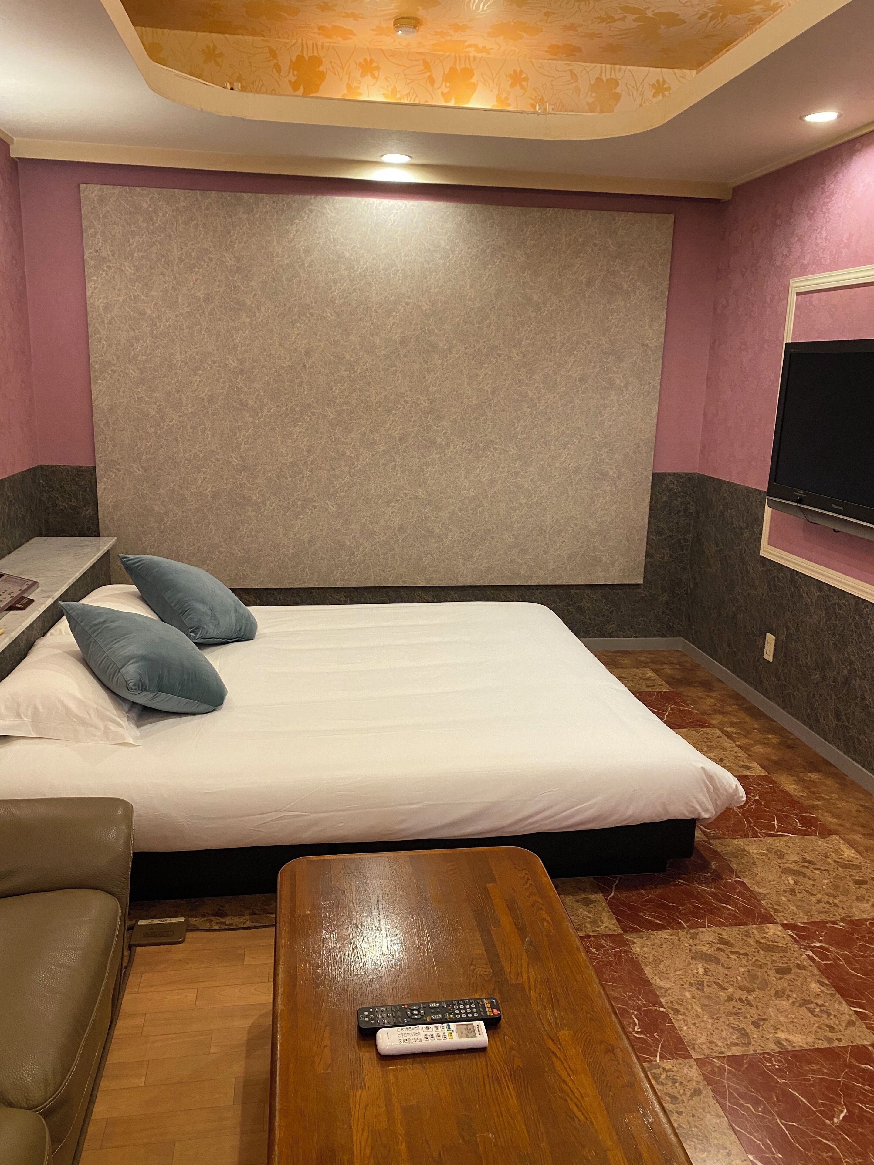Ein Hotelzimmer mit einem großen Bett, einem an der Wand montierten Fernseher, einem Holztisch und einer Fernbedienung auf dem Tisch.