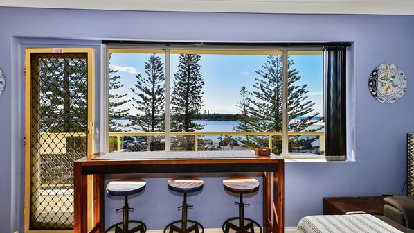 Ein Zimmer mit großem Fenster mit Blick auf einen See und Bäume, ein Holztisch mit Barhocker und eine Couch.