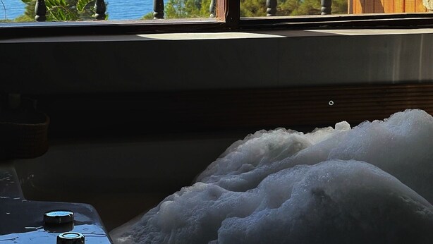 A window with a view of a lake and mountains, a wooden deck, and a bathtub filled with bubbles. - Faralia Hotel (Fethiye)