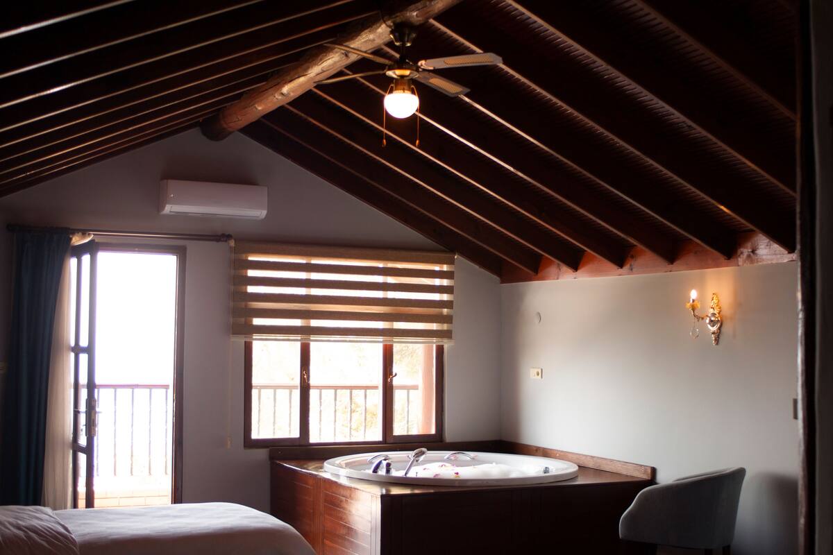 A room with a wooden ceiling, a large window with blinds, a round bathtub, a bed, and a chair.
