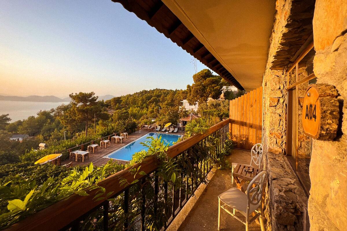 A balcony with a stone wall, a wooden railing, and a view of a pool area with lounge chairs and umbrellas.