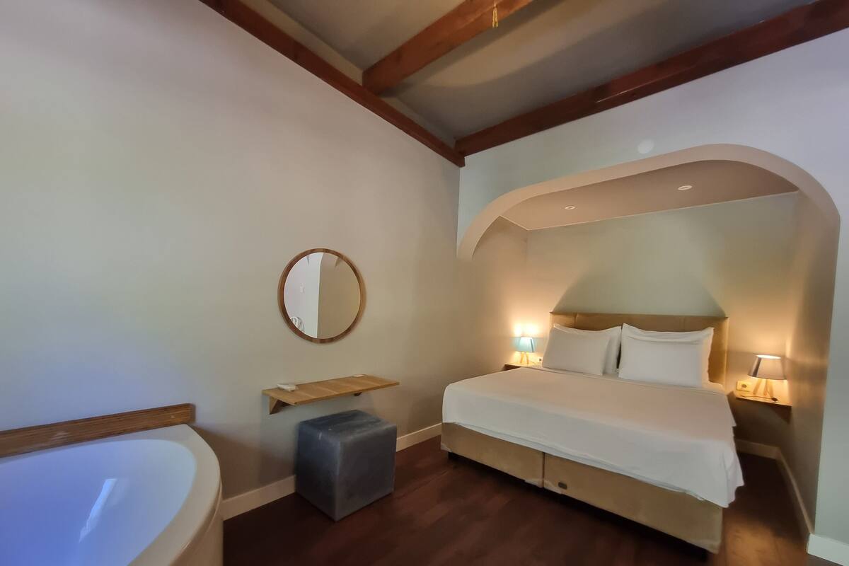 A bedroom with a large bed, a bedside table, a nightstand, a round mirror, a small stool, and a bathtub.