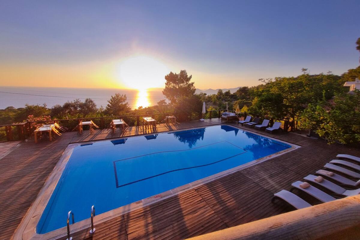 A rectangular swimming pool with a wooden deck, surrounded by lounge chairs and tables, overlooking a scenic view of the sea and mountains during sunset.