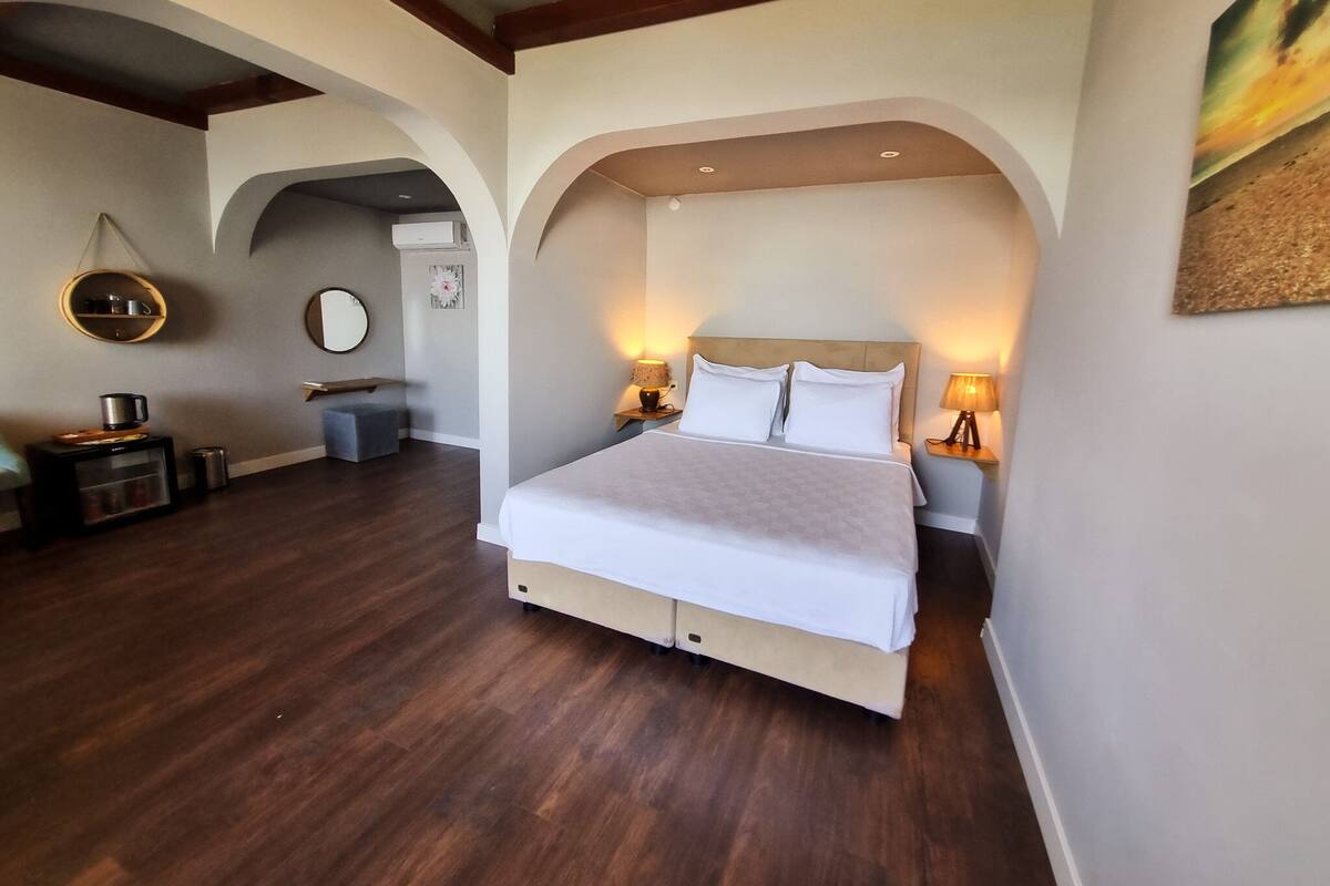 A hotel room with a large bed, two bedside tables with lamps, a round mirror, a small seating area, and a wooden floor.