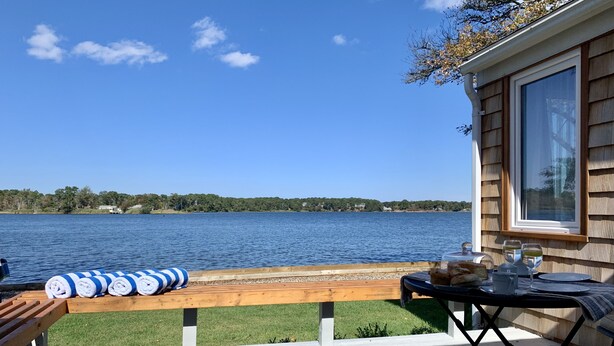A deck with a view of a lake, a wooden bench, and a table with bread and drinks. - Cape Code Waterfront Cottage (Dennis Port)