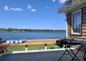 A deck with a view of a lake, a wooden bench, and a table with bread and drinks. - Cape Code Waterfront Cottage (Dennis Port)