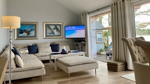 A modern living room with a sofa, ottoman, and a TV. There are framed pictures on the wall, a lamp, and a view of the outdoors. - Cape Code Waterfront Cottage (Dennis Port)