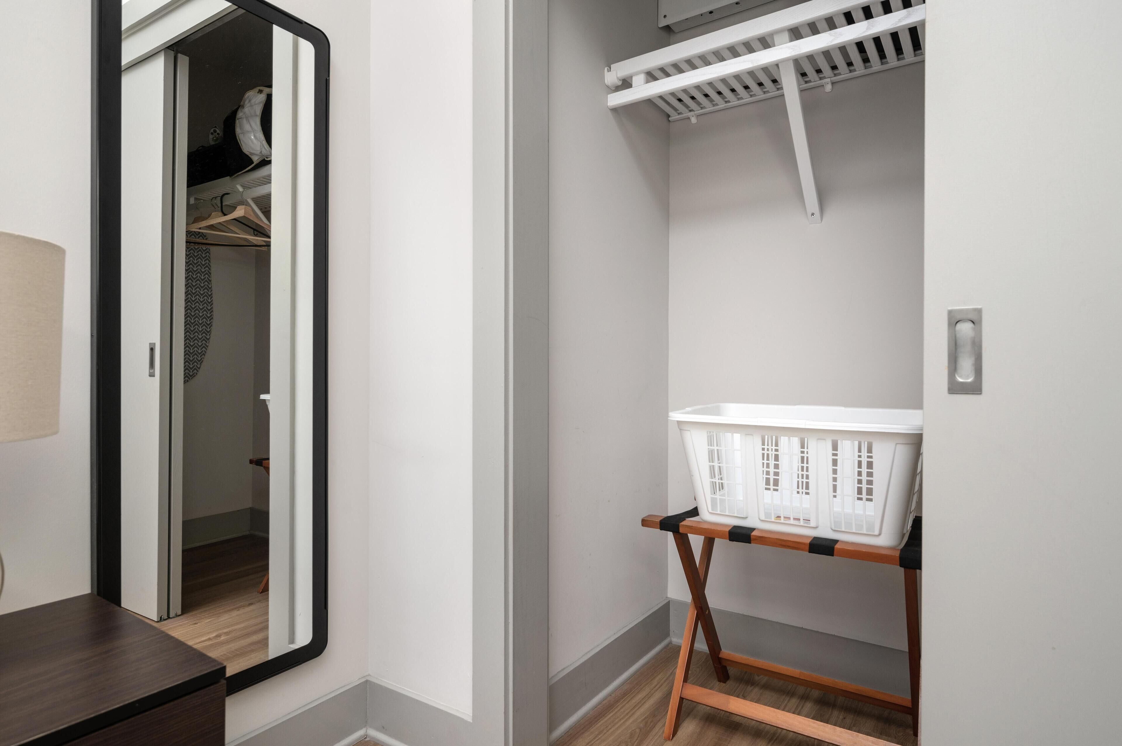 Quarto com piso de madeira, parede branca, espelho, mesa de cabeceira de madeira, cesto de roupa branca sobre suporte de madeira e ventilador de teto branco.