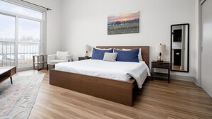 Un dormitorio moderno con una cama grande, un sillón blanco, una mesita, un espejo y un cuadro con un atardecer sobre un campo.