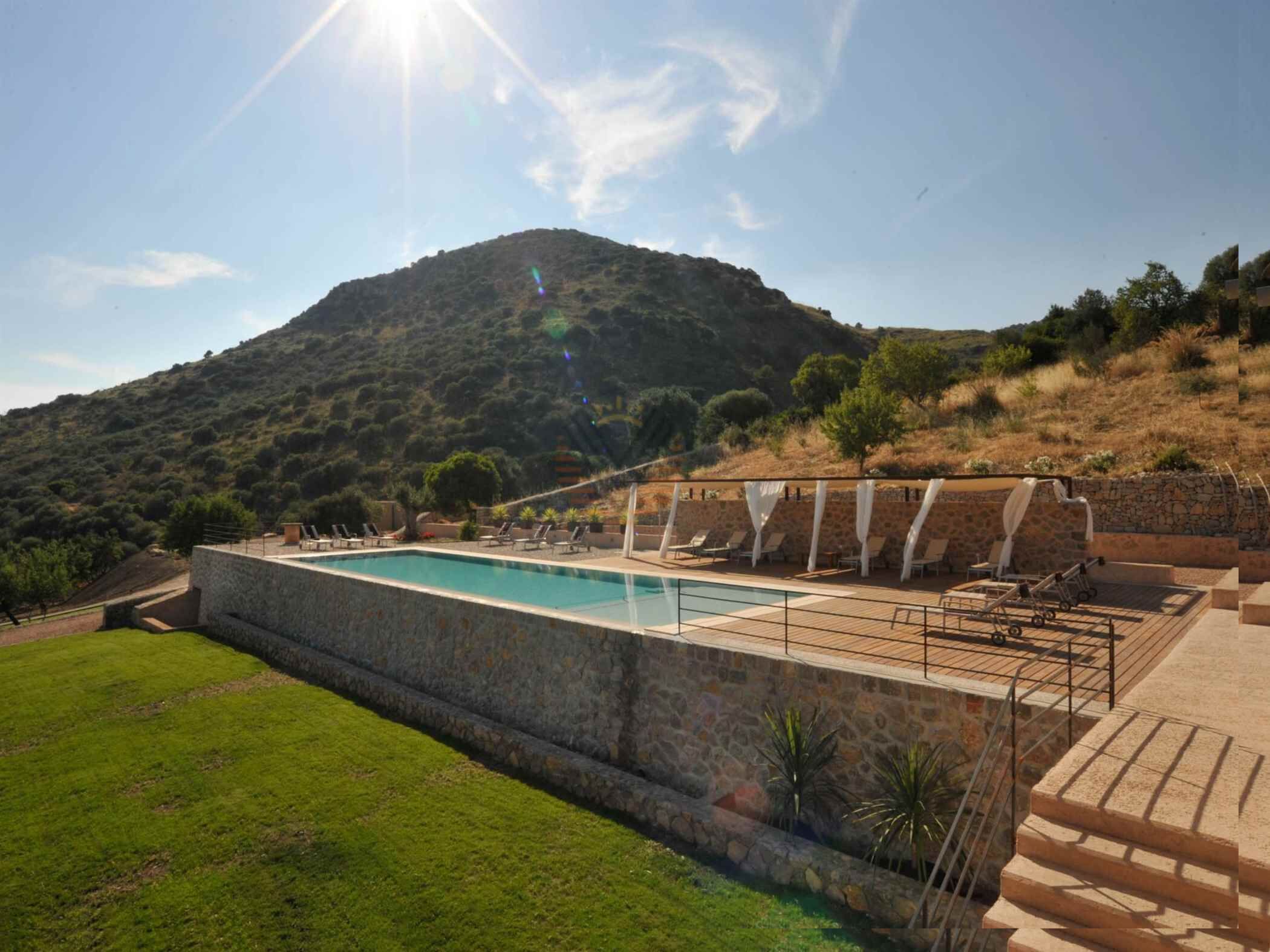 Une piscine bordée d’un mur en pierre, entourée de chaises longues et offrant une vue sur une colline.