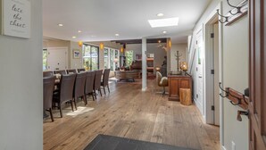 Un grand espace dédié à la salle à manger, avec une longue table et des chaises, un coin salon confortable avec une table ronde, et une cuisine équipée de meubles en bois et d’un évier.
