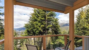 Un balcón con dos sillas, baranda de madera y vista a un bosque de coníferas y montañas.