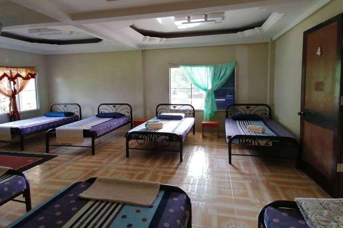 A dormitory room with four single beds, a small wooden bench, and a window with a green curtain.