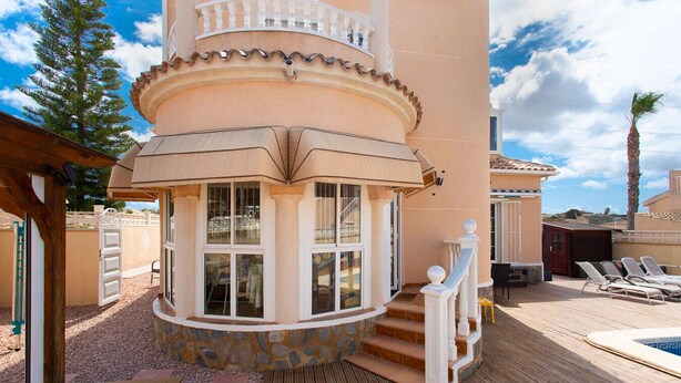 A corner of a building with a stone base, a balcony with white railings, and a pool area with lounge chairs.