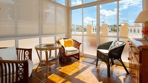 A sunlit room with large windows, wicker furniture, and a view of a coastal area.