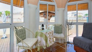 A sunlit room with two wicker chairs, a table with a floral tablecloth, and a grey sofa.