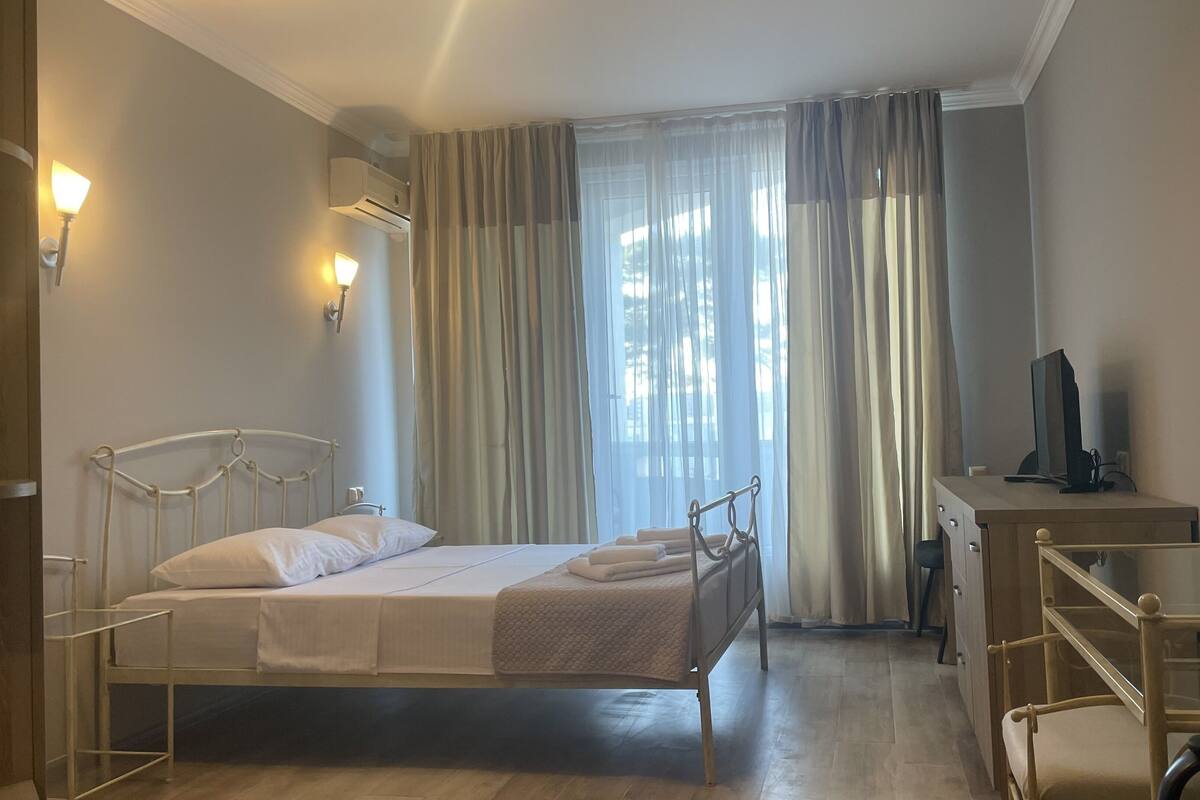 A hotel room with a single bed, a desk with a computer, a mirror, and a window with sheer curtains.